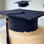 Elegant graduation cake topped with a realistic fondant graduation cap featuring a tassel and button, with additional caps in the background